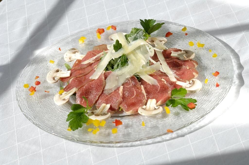 Un plato de vidrio transparente con carne en rodajas, champiñones y guarniciones sobre un mantel blanco.