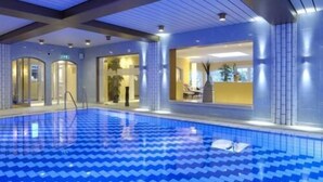 An indoor swimming pool with a blue chevron pattern on the floor, surrounded by walls with a modern design and glass doors leading to a dining area. - Posthotel Rattenberg (Rattenberg)