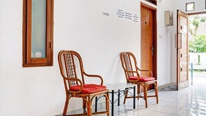 Un couloir avec deux fauteuils en osier dotés de coussins rouges, un sol carrelé et une porte en bois sur laquelle est apposée une plaque.