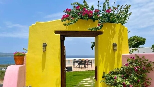 Uma porta amarela dá para um pátio com vista para o mar, móveis de exterior e plantas em vasos.