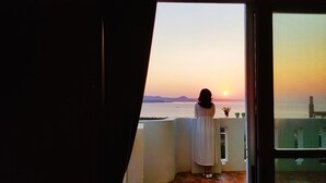 Uma pessoa parada junto a uma janela com vista para o pôr do sol sobre o mar, com varanda e vista de colinas distantes.