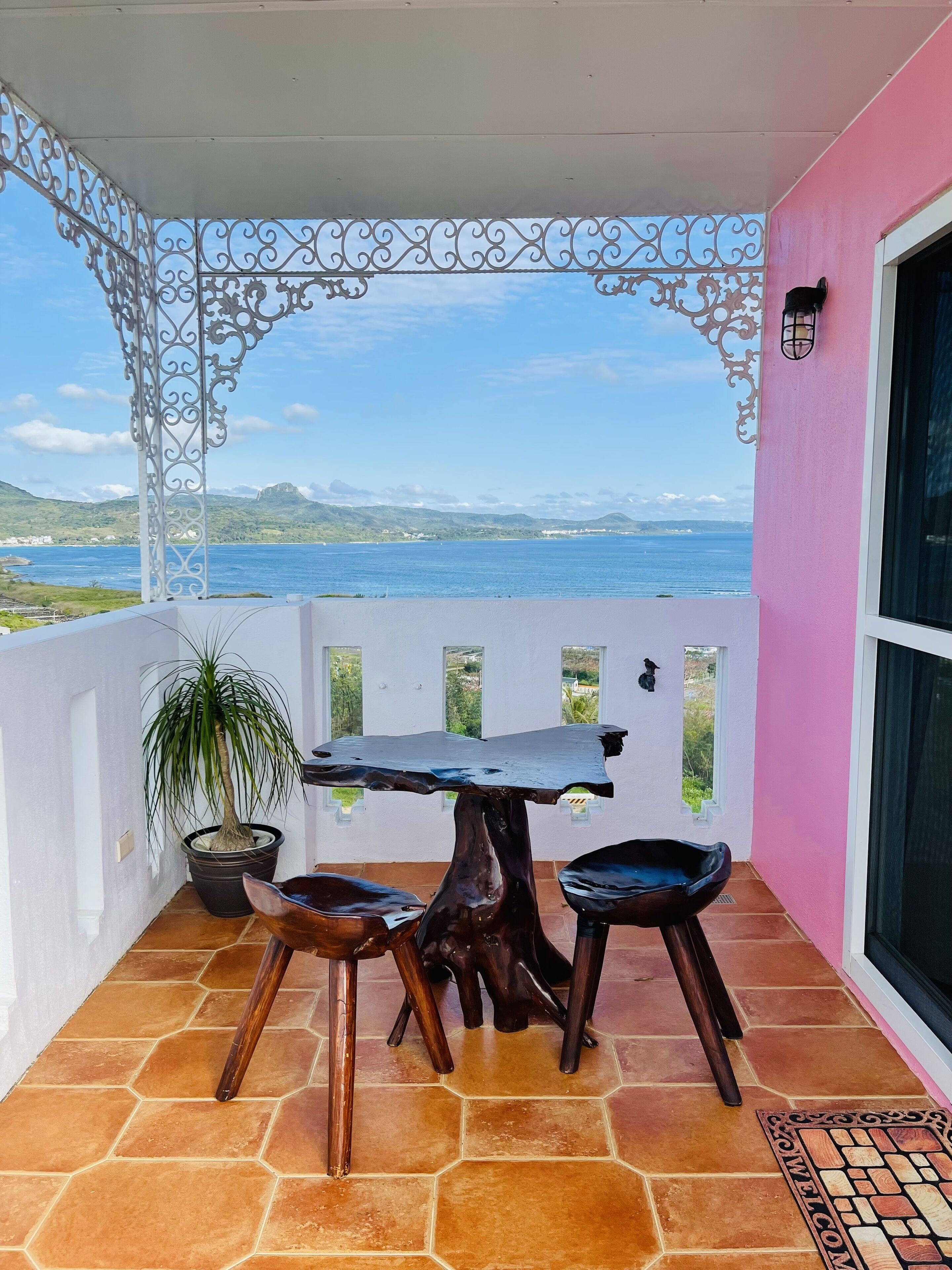 A balcony with a table and chairs, a potted plant, and a view of the sea and hills.