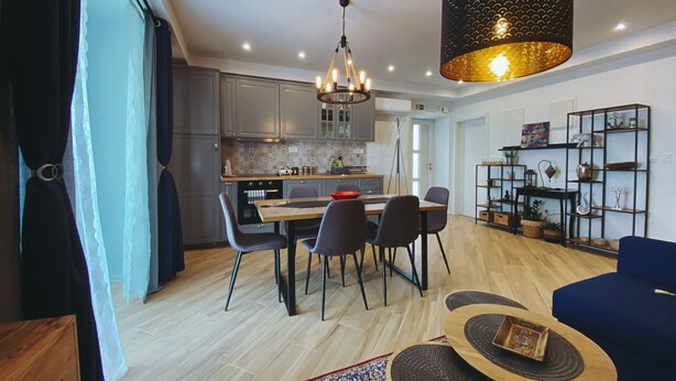 A modern kitchen with grey cabinets, a dining area with a wooden table and chairs, a blue sofa, and a decorative shelf with various items. - Villa Marghareta 2 (Dubrovnik)