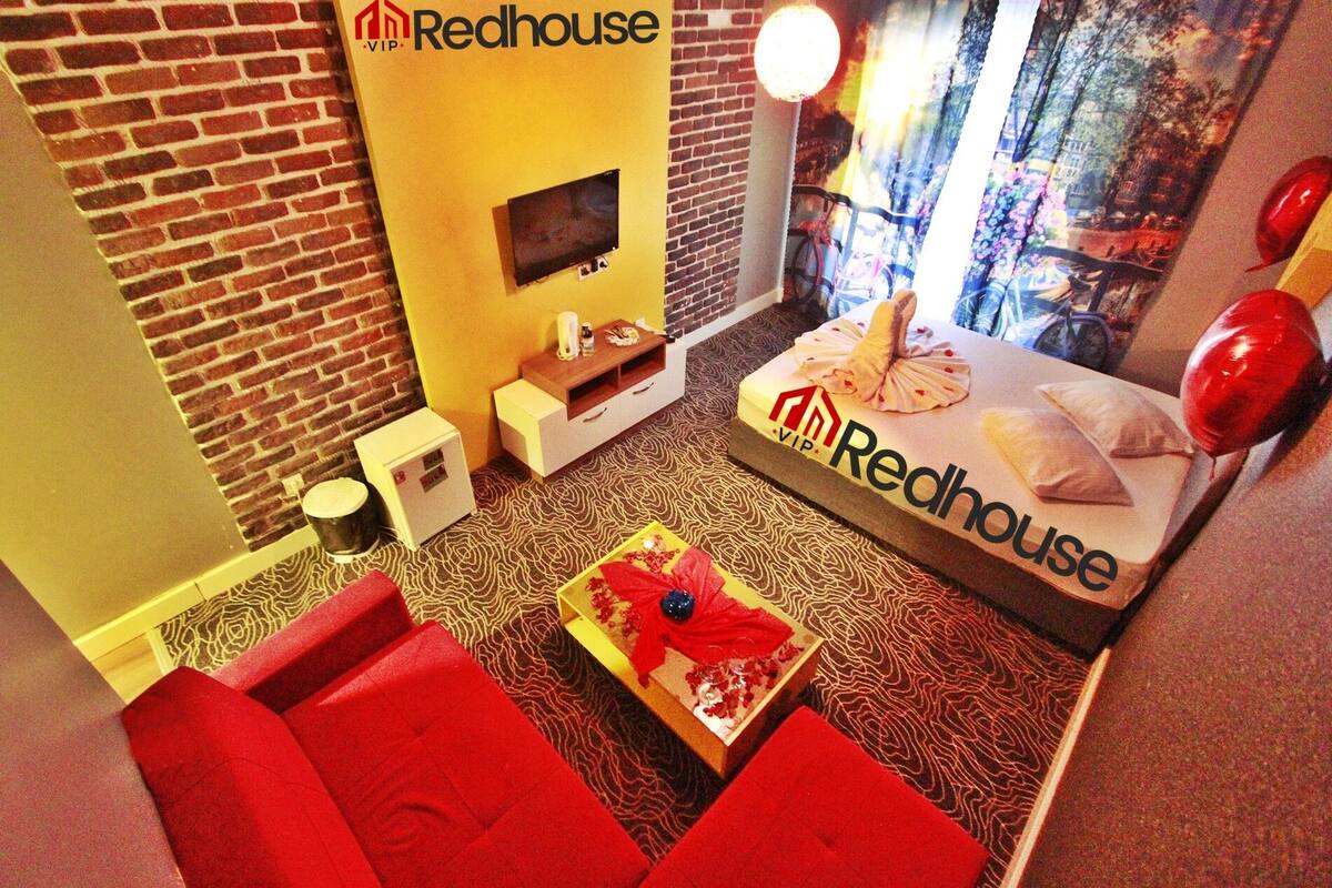 A Redhouse VIP room with a brick wall, a flat-screen TV, a bed with Redhouse branding, a red sofa, a patterned carpet, and a small table with a decorative item.