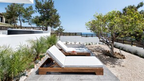 Ein moderner Außenbereich mit einer Lounge-Ecke, bestehend aus zwei Holzliegen und einem kleinen Tisch, umgeben von einem angelegten Garten mit verschiedenen Pflanzen und Blick auf das Meer.