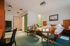 A hotel room with a bed, two wooden chairs, a small table with a vase of flowers, a painting on the wall, and a desk with a chair.