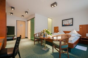 Habitación de hotel con una cama, dos sillas de madera, una mesita con un jarrón de flores, un cuadro en la pared y un escritorio con una silla.