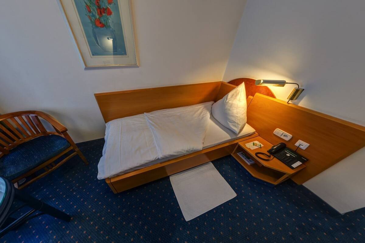 A single bed with white linens, a wooden headboard, a bedside table with a lamp, a chair, and a small desk with a telephone.