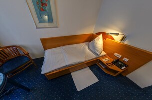 Una cama individual con ropa de cama blanca, cabecera de madera, mesita de noche con lámpara, una silla y un escritorio pequeño con teléfono.