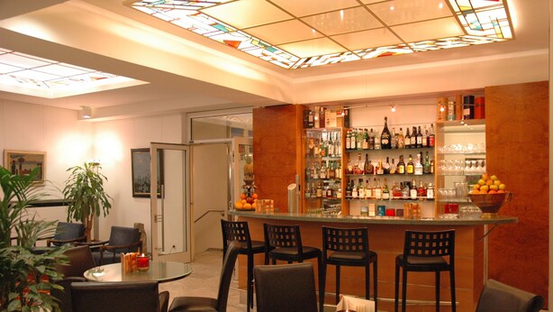 Zona de bar con una mesa de vidrio, taburetes y una variedad de botellas y vasos en estanterías.