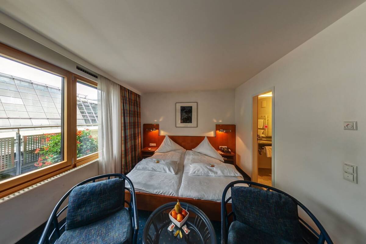 A hotel room with a bed, two chairs, a small table with fruit, a window with curtains, and a bathroom visible through an open door.