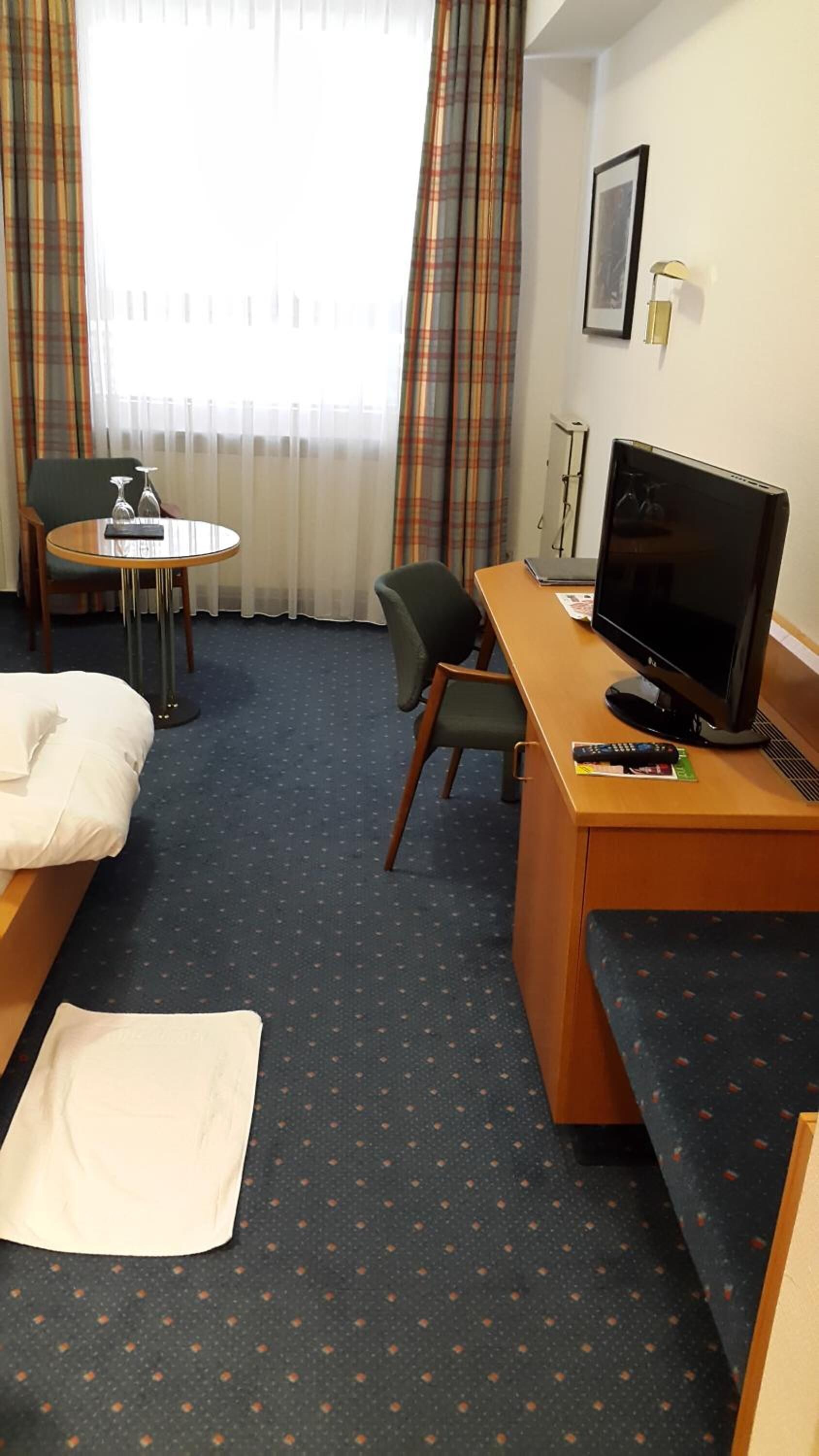 Habitación de hotel con una cama, un escritorio con televisor, una silla, una mesita con un vaso y una ventana con cortinas.