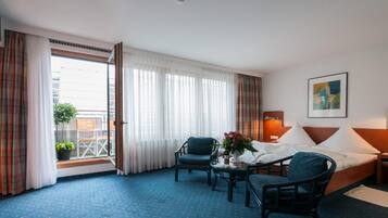A hotel room with a bed, two chairs, a small table with a vase of flowers, a nightstand, and a painting on the wall.