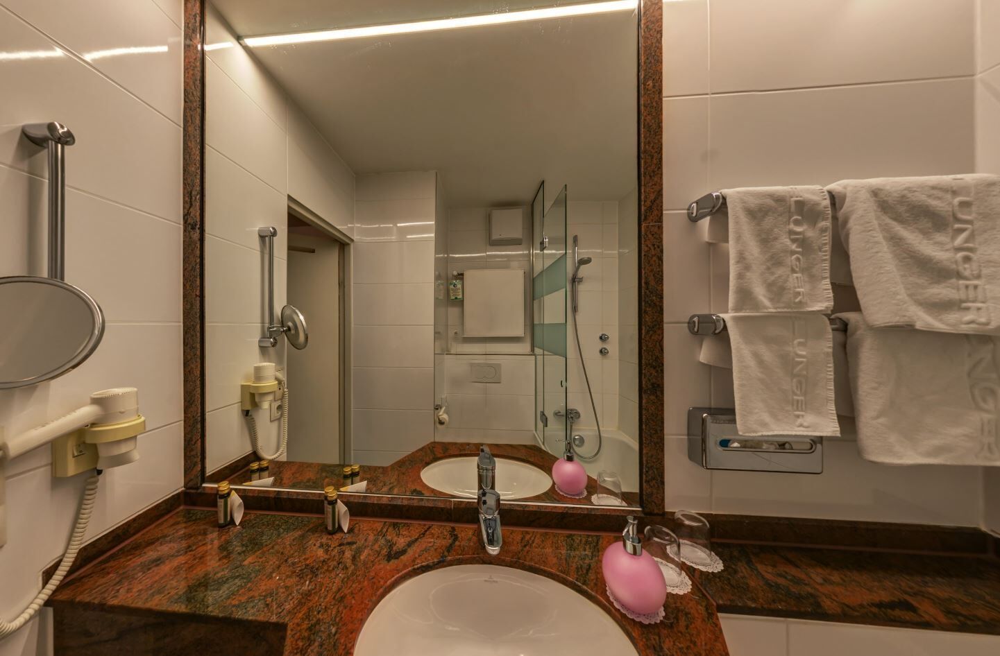 A hotel bathroom with a large mirror, two sinks, a shower, and a towel rack with white towels.