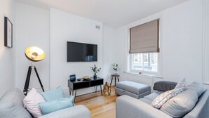 A modern living room with grey sofas, a black entertainment unit, a floor lamp, and a large window with a beige blind. - Deluxe 4 BR Oxford Circus Apartment with Terrace (London)