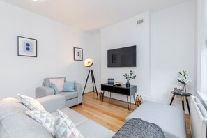 A modern living room with a grey sofa, a small table with a lamp, a TV mounted on the wall, and a small plant on a side table. - Deluxe 4 BR Oxford Circus Apartment with Terrace (London)