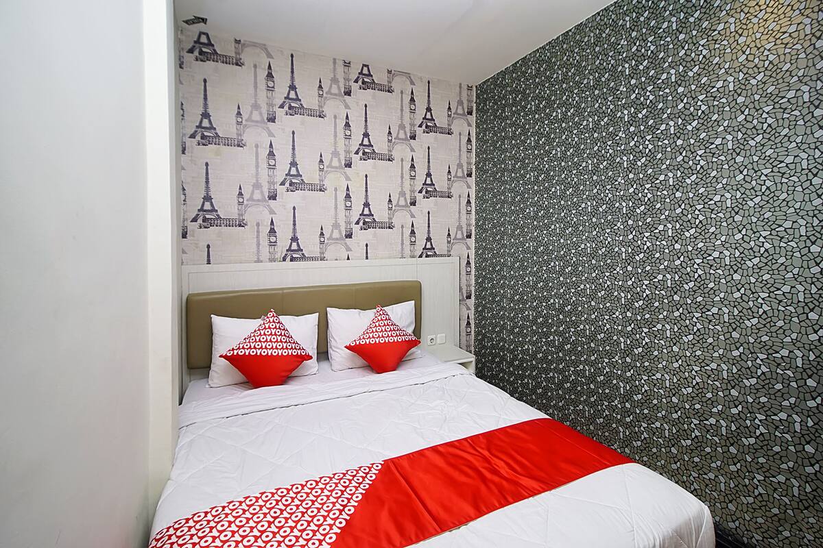 A bed with a white and red bedspread, two red pillows, and a headboard. The wall behind the bed has a pattern of the Eiffel Tower and a mosaic design.