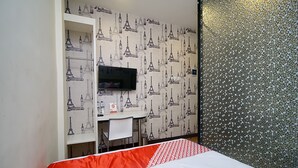 Une chambre d’hôtel avec un lit, un téléviseur fixé au mur et orné d’un papier peint à motifs de la Tour Eiffel, un petit bureau avec une chaise et un mur décoré de motifs en mosaïque.