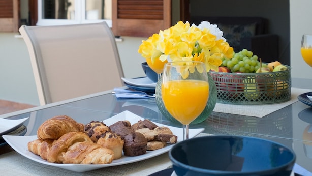 Une table de petit-déjeuner avec des croissants, des biscuits, un panier de fruits, un verre de jus d’orange et un vase rempli de fleurs.