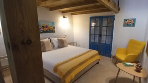 Un dormitorio con una cama, un sillón amarillo, una mesita, una puerta azul y un cuadro en la pared.