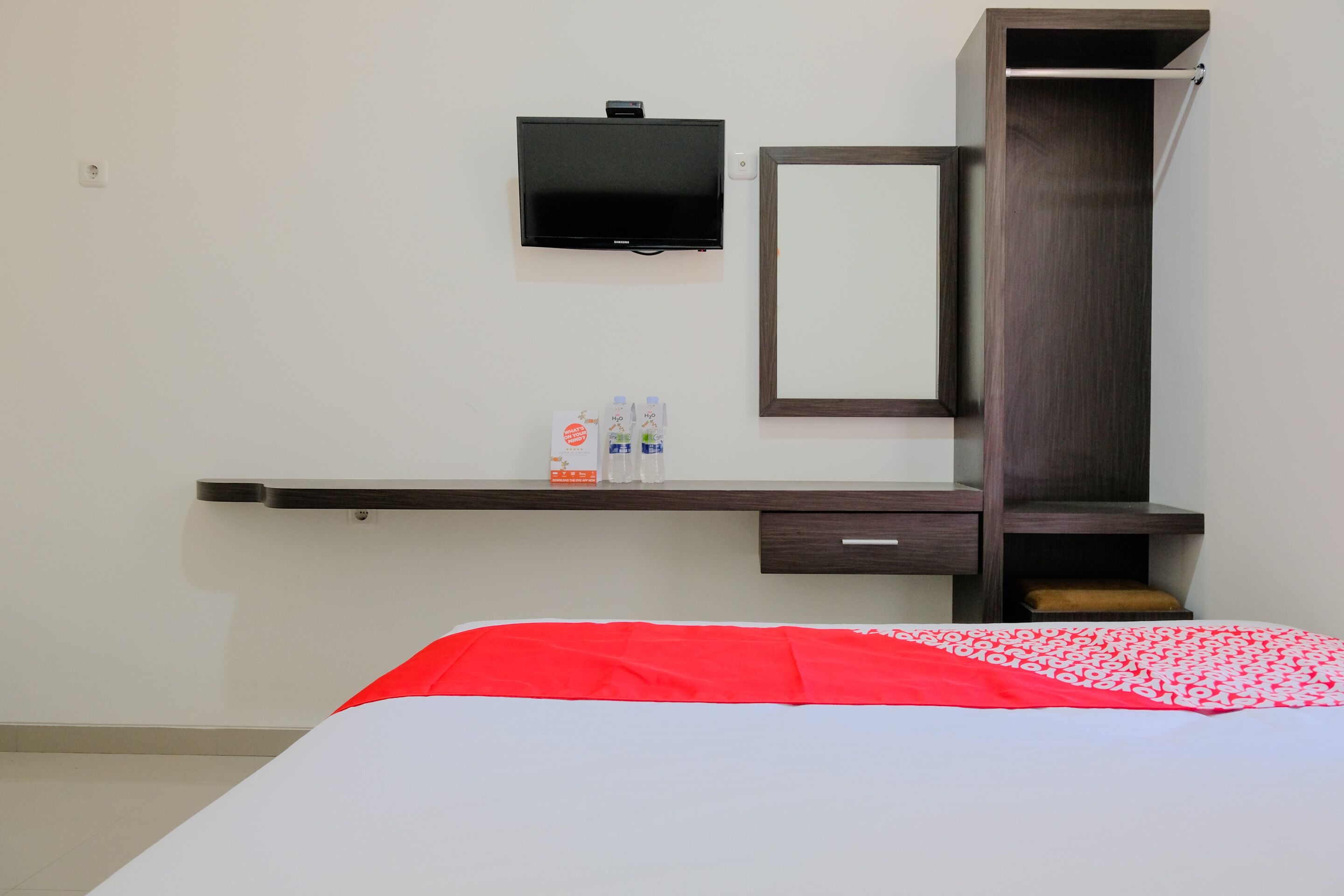 A hotel room with a bed, a TV mounted on the wall, a desk with a mirror, and a shelf with toiletries.