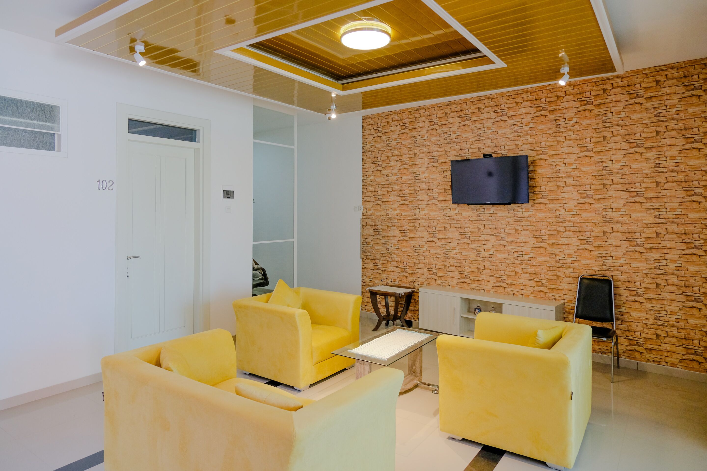 A modern room with yellow sofas, a flat-screen TV mounted on a brick wall, and a wooden ceiling.