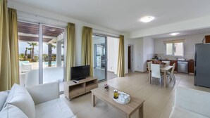 A modern living room with a flat-screen TV, a coffee table, and a dining area with a kitchen in the background. - A Truly Spectacular First-Line Sea Front Villa in the beautiful village of Pervolia (Perivolia)