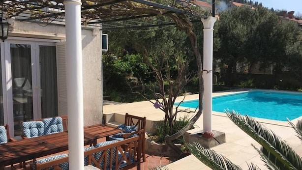 A poolside area with a wooden table and chairs, a pergola, and a view of a mountain and buildings in the background. - Villa Marghareta - Deluxe App Marghareta (Dubrovnik)