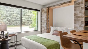 A modern hotel room with a large window, a bed with a green throw, a dining table, and a built-in wardrobe. - Downtown Aspen Studio | Walk to Gondola & Slopes (Aspen)