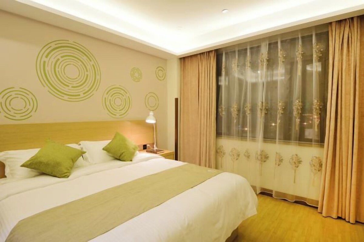 Una habitación de hotel con una cama grande, una mesita de noche con lámpara, una ventana con cortinas translúcidas y detalles decorativos en la pared.