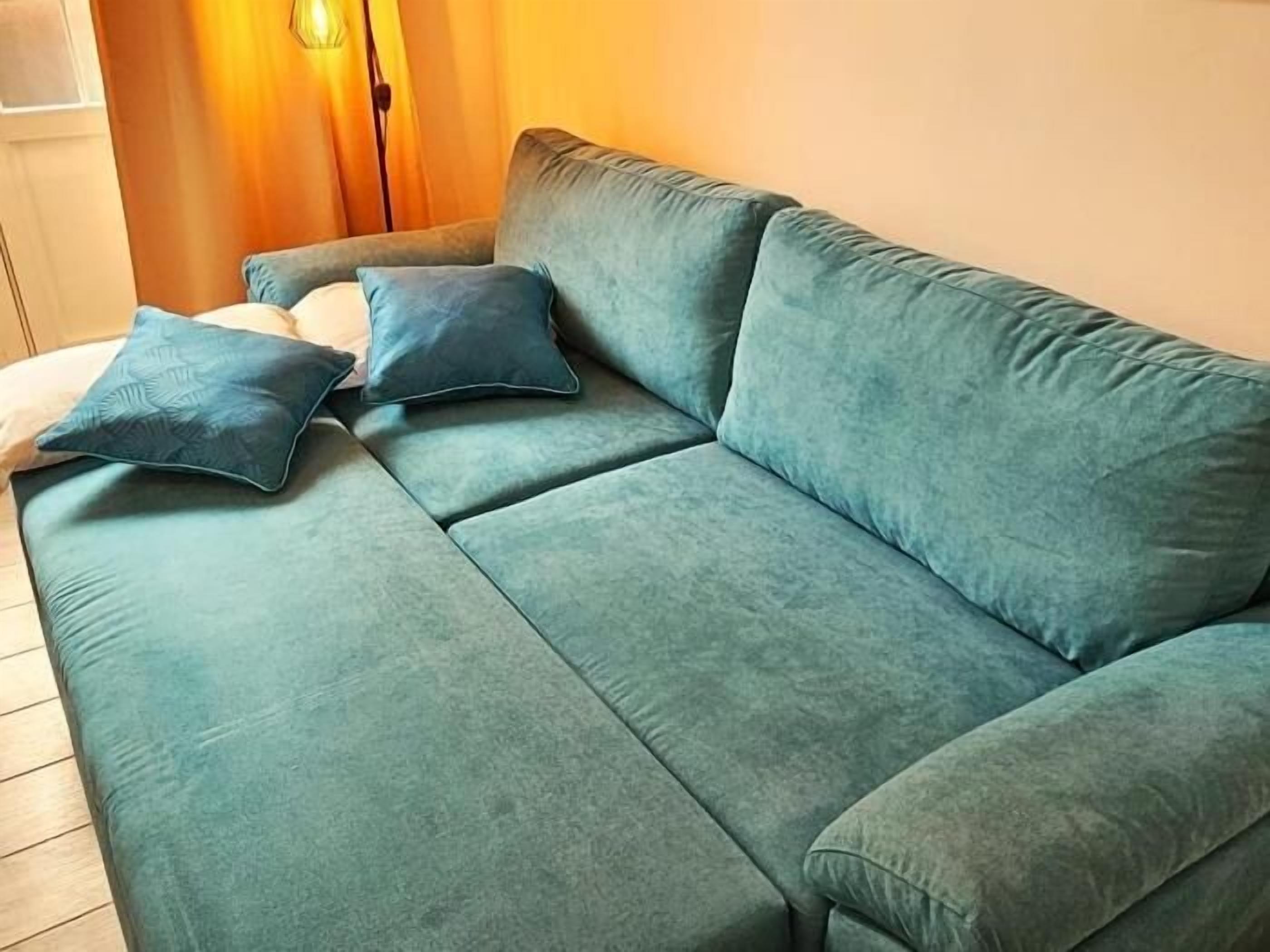 A teal sofa with two pillows, a wooden floor, and a curtain in the background.