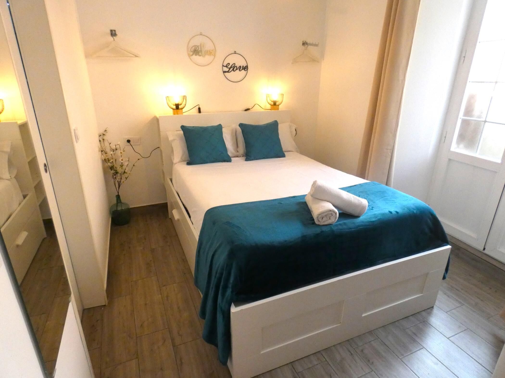 A neatly made bed with a blue bedspread and pillows, a white bedside table, and a wall-mounted light fixture.