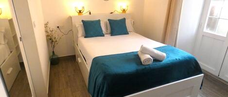 A neatly made bed with a blue bedspread and pillows, a white bedside table, and a wall-mounted light fixture.
