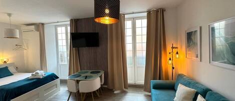 A modern hotel room with a bed, a sofa, a dining table with chairs, a TV mounted on the wall, and a large window with curtains.
