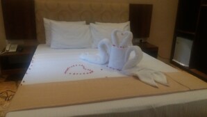 A neatly made bed with a swan and heart-shaped towel arrangement, a bedside phone, and a nightstand with a lamp. - Hotel Asliya Regency (Mawathagama)
