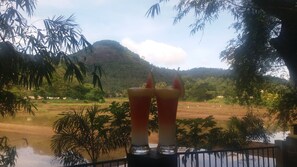 A view of a river with a mountain in the background and two glasses of red beverage on a table. - Hotel Asliya Regency (Mawathagama)