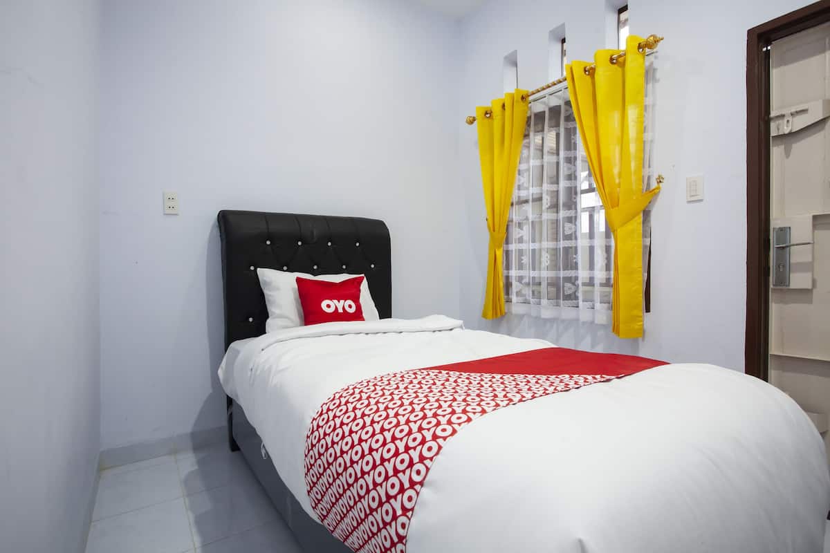 A bed with a black headboard, a white and red patterned bedspread, and a red pillow with "OYO" text. Yellow curtains are partially drawn, and a window with a white curtain is visible.