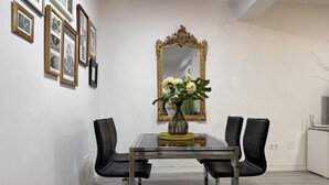 Een eetgedeelte met een glazen tafel, zwarte stoelen, een spiegel met gouden lijst en een vaas met bloemen.