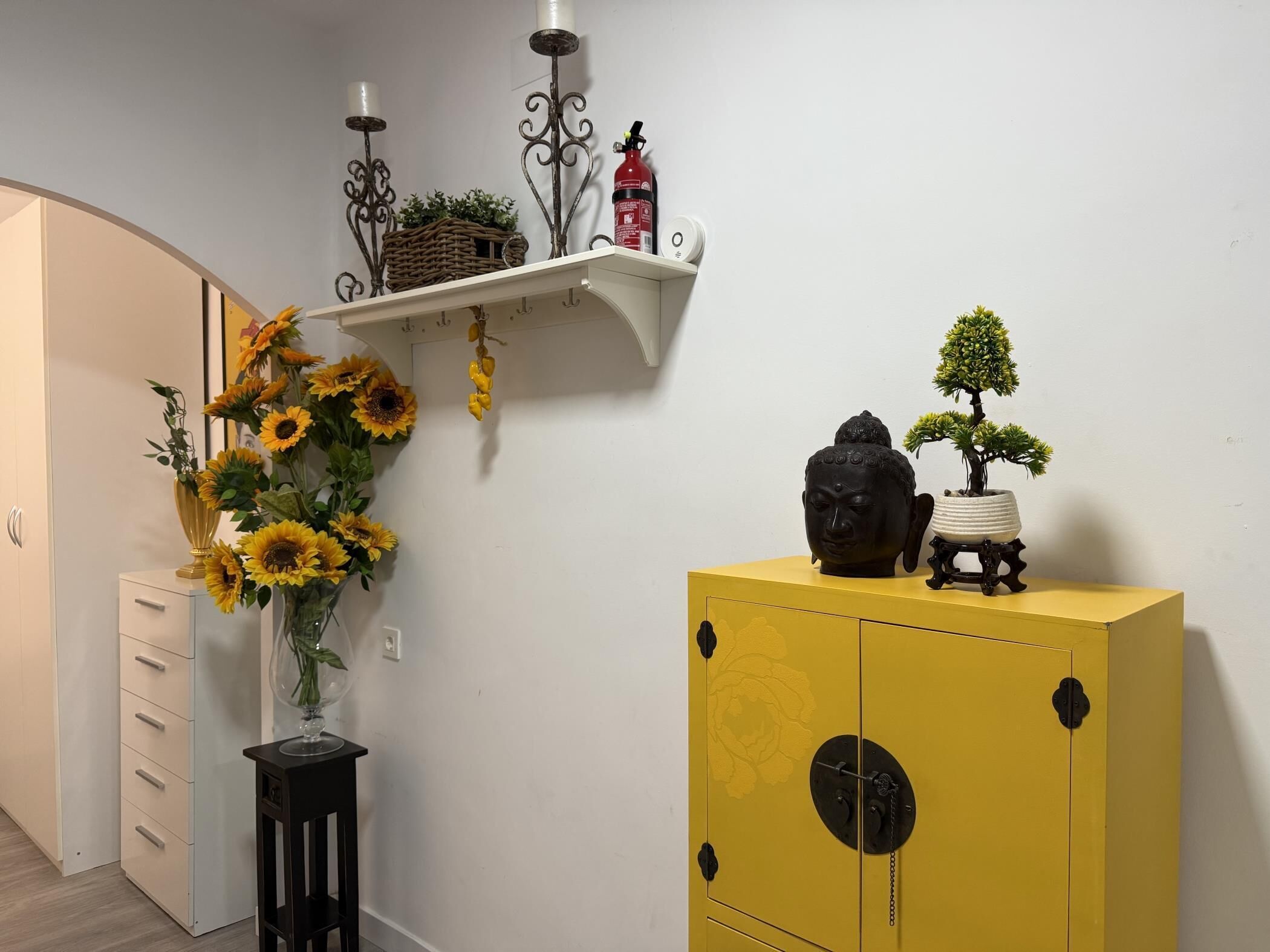 Ein Flur mit einer weißen Wand, einem gelben Schrank mit Buddha-Statue, einem kleinen Regal mit Kerzen und einem Feuerlöscher sowie einer Vase mit Sonnenblumen.