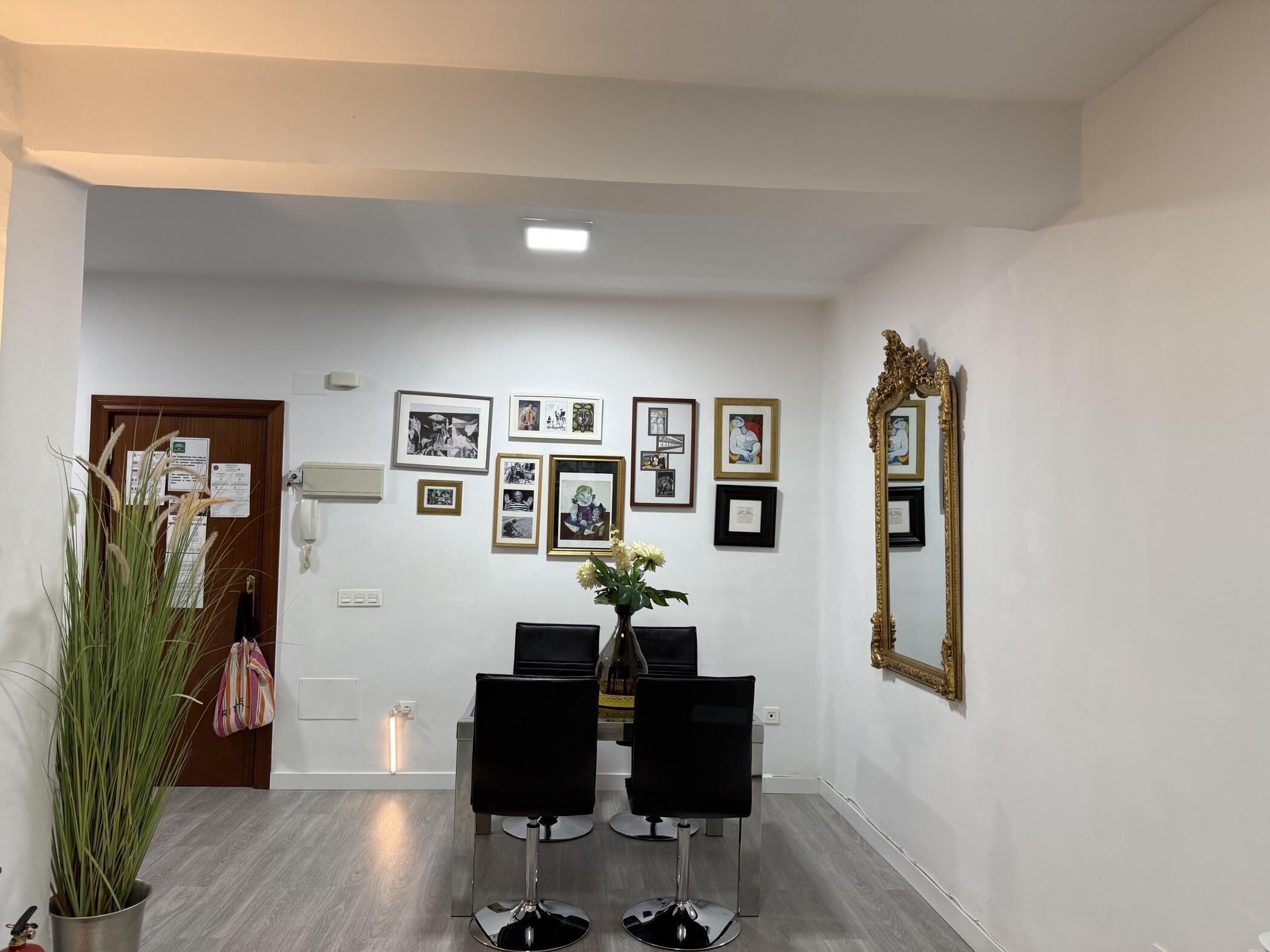Um corredor com área de refeições, com duas cadeiras pretas, um vaso de flores e uma parede decorada com obras de arte emolduradas.