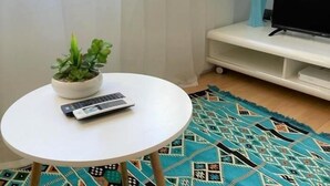 Una pequeña mesa redonda con una planta en maceta y un control remoto, un tapete estampado, un televisor sobre un mueble bajo y una ventana con cortinas translúcidas.