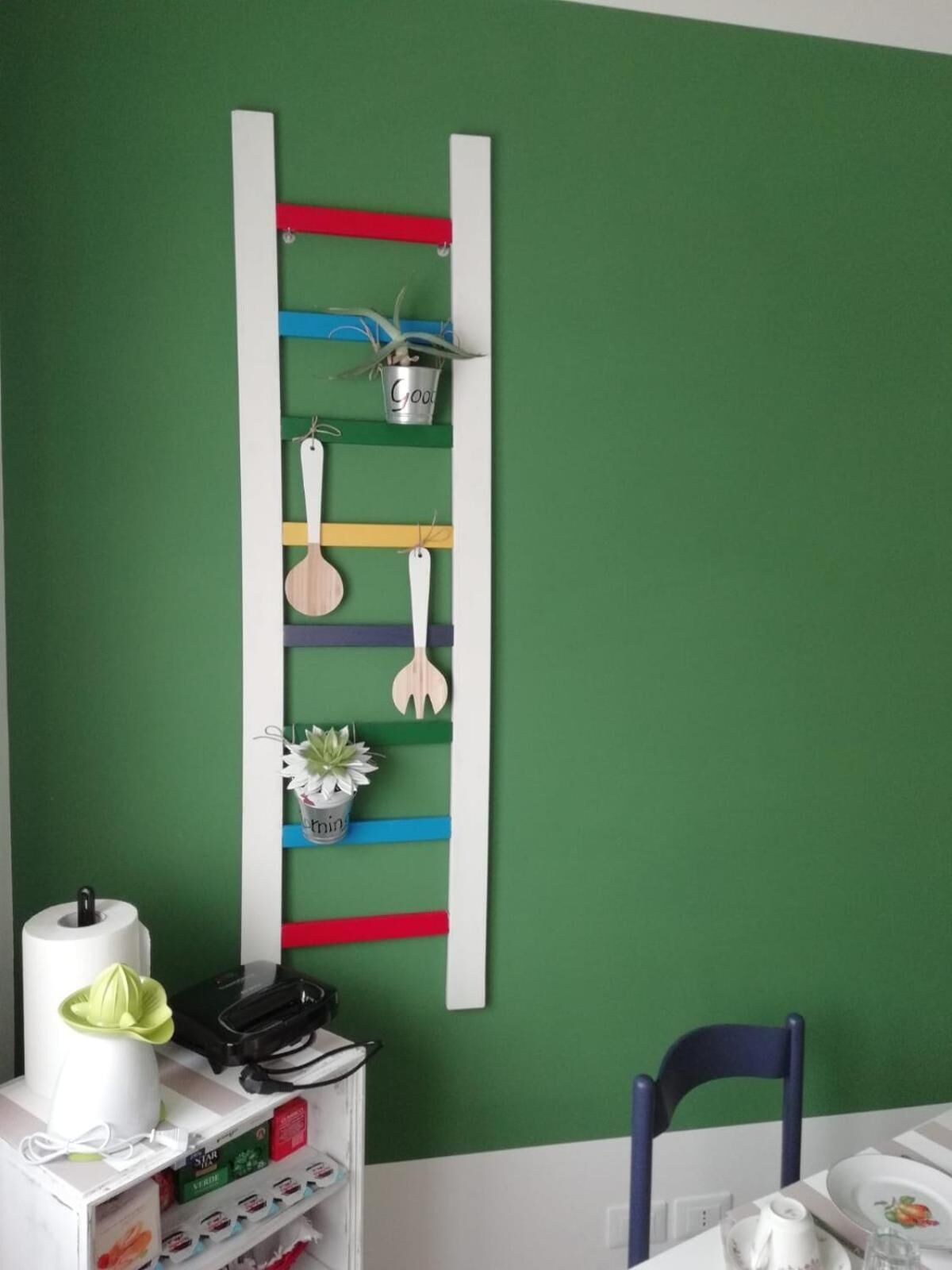 Una cocina con una pared verde, un estante blanco con utensilios de cocina, una silla azul y una mesa blanca.