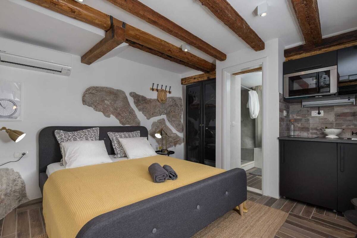 A bedroom with a bed, bedside lamps, a stone wall, and a kitchen area with a microwave.