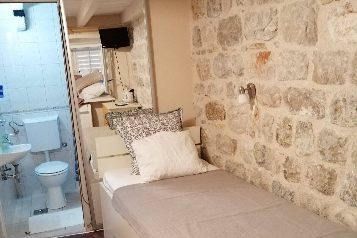A bedroom with a stone wall, a bed with a beige bedspread, a bedside table with a lamp, and a view into the bathroom with a toilet and sink.