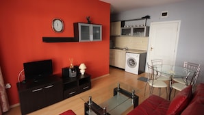Un espacio habitable compacto con una pared roja, un televisor de pantalla plana, una mesa de centro de vidrio, un sofá rojo, una zona de cocina con lavadora y una mesa de comedor con sillas.