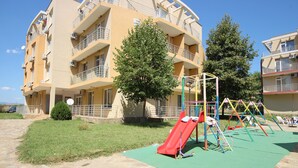 Un parque infantil con un tobogán rojo y columpios, rodeado por un tapete verde, frente a un edificio de varios pisos con balcones y unidades de aire acondicionado.