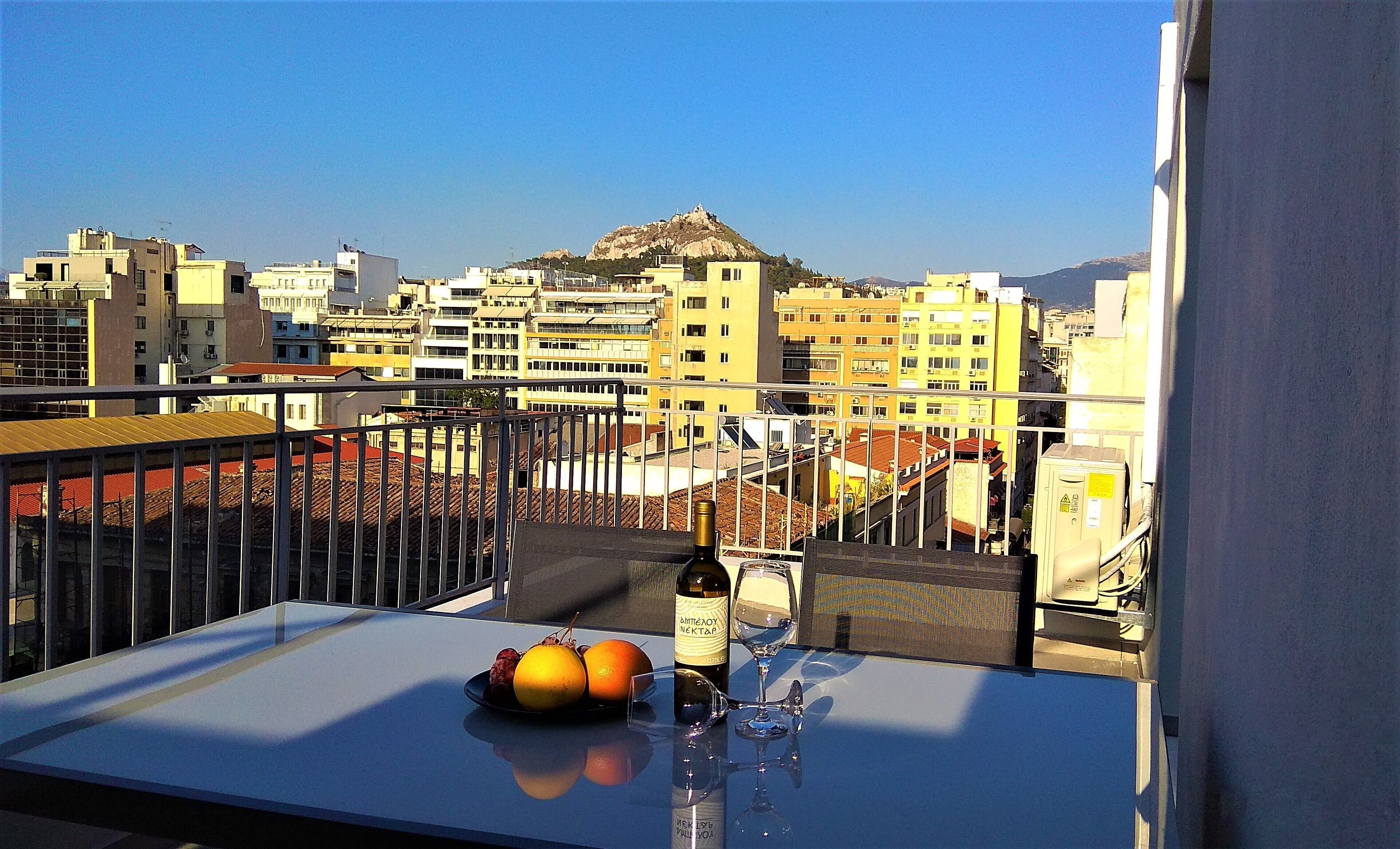 A balcony with a table, a bottle of wine, and a glass, overlooking a cityscape with buildings and a mountain.