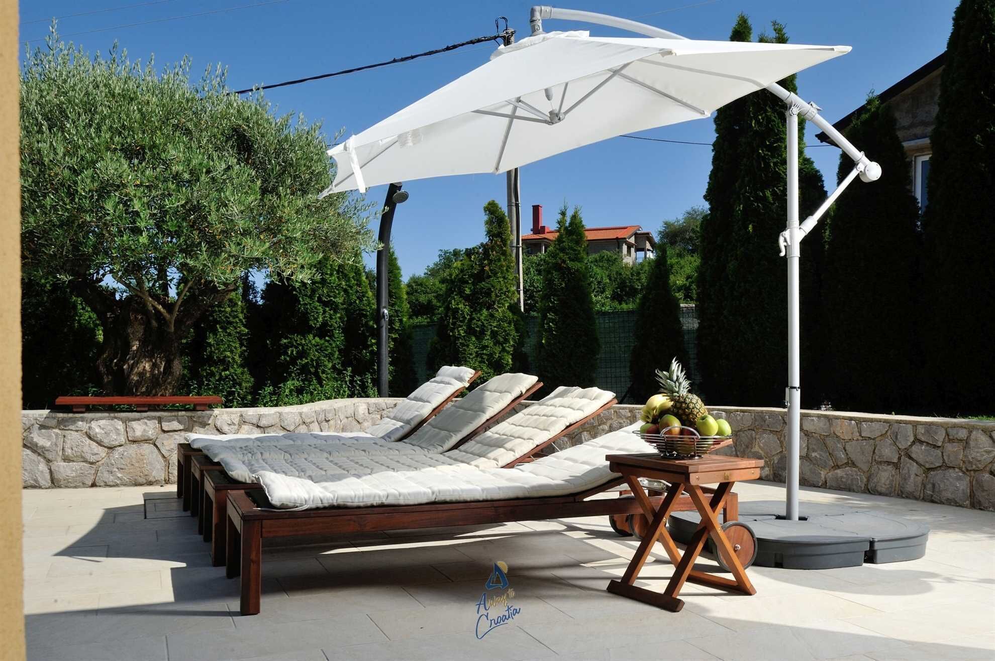 A sunbed with a white umbrella, a wooden table with fruit, and a stone wall.