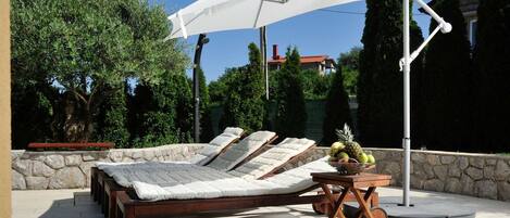 A sunbed with a white umbrella, a wooden table with fruit, and a stone wall.