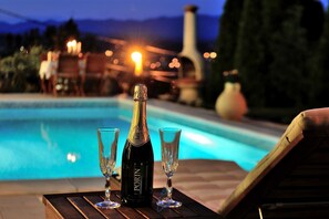 Una botella de champán PORIN y dos copas sobre una mesa de madera junto a una piscina, con vistas a la montaña al atardecer.